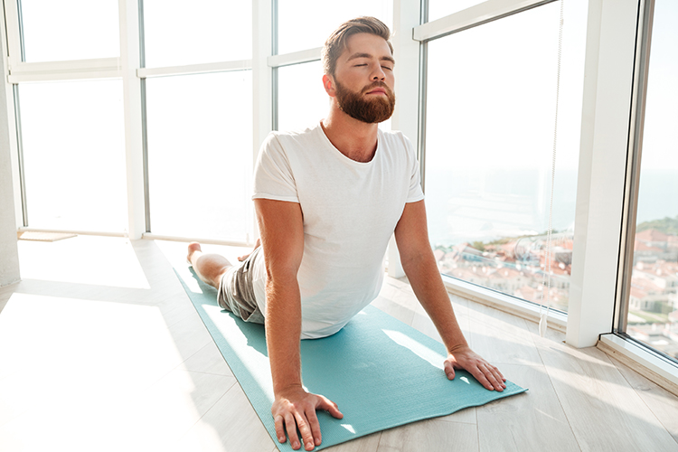 Man with the Dolphin chronotype performing a stretching exercise in a yoga pose on a mat by a large window in the morning
