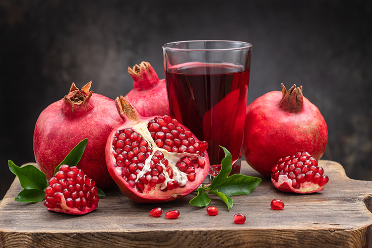 Pomegranate drink in a clear glass and juicy open pomegranate fruits with pomegranate leaves on a wooden stand.