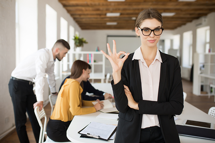 Young calm and smiling businesswoman in glasses and shirt showing an “OK” gesture while spending time in the office with colleagues